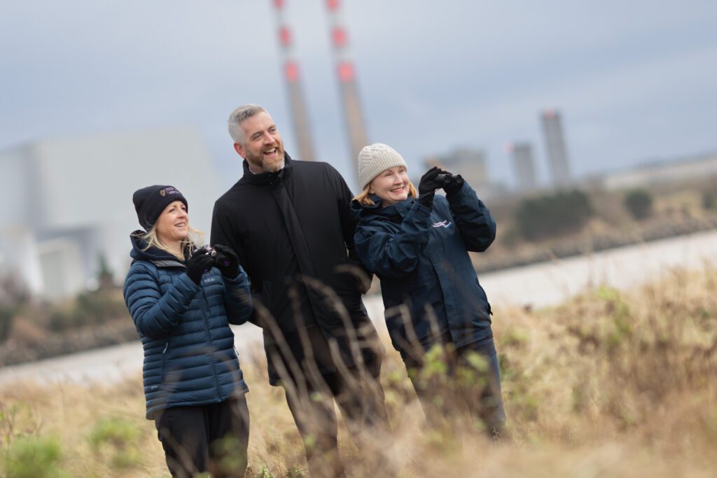 Two women and a man bird-watching on the coast of Dublin. Dressed in outdoor clothes and hat, Deirdre Lynn of the National Parks and wildlife service is holding binoculars. Standing in middle of group with a big outdoor coat is Minister Christopher O'Sullivan, also standing next to him with binoculars which she is holding aloft is Moninne Griffith of the Community Foundation. All three are standing in coastal grasses about knee high.